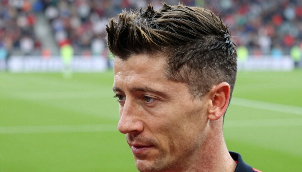 A close-up portrait of Robert Lewandowski showcasing his iconic hairstyle, featuring a precise undercut with longer, styled hair on top, slightly tousled for a modern and dynamic look. The foreground captures the vibrant glossy texture of his hair, reflecting subtle highlights under soft, natural lighting. In the middle ground, include a blurred football pitch to emphasize his connection to the sport, with faint crowd silhouettes in the background. The image is taken from a slightly elevated angle to highlight the hairstyle, creating an energetic and professional atmosphere suitable for illustrating haircut techniques. The setting conveys a sense of dedication and style, resonating with fans and aspiring footballers.